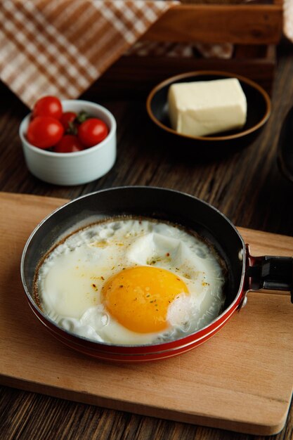 Side view of fried egg in frying pan on cutting board with tomatoes butter on wooden background