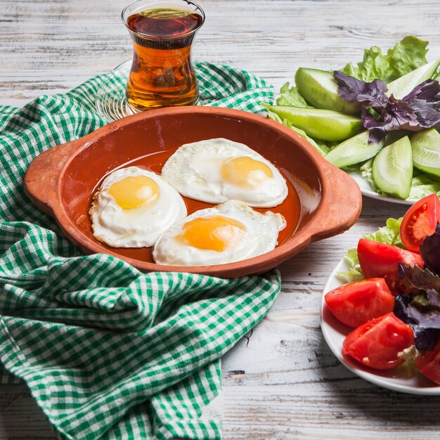 Вид сбоку жареных яиц с нарезанными помидорами и нарезанными огурцами и стаканом чая в глиняной тарелке