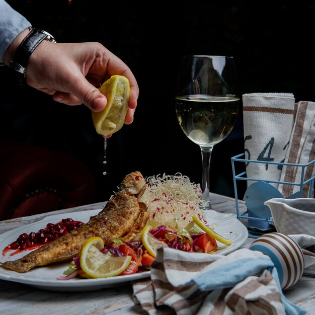 Side view fried fish with glass of wine and lemon and human hand in white plate