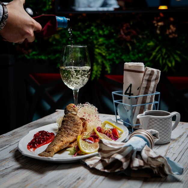 Side view fried fish with glass of wine and wine bottle and human hand in white plate