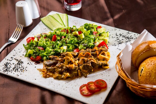 Side view fried meat with mushrooms in sauce with vegetable salad and slices of tomato and cucumber