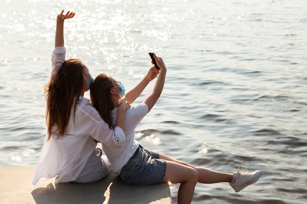 Side view of friends with medical masks taking selfie by the lake