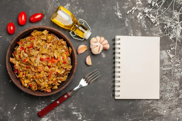 Side view from afar appetizing dish appetizing dish next to the fork bottle of oil garlic tomatoes and white notebook on the dark table