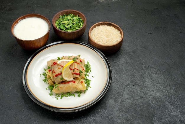 Side view from afar appetizing dish appetizing stuffed cabbage with herbs lemon and sauce on white plate and plates with sour cream herbs and rice in the center of black table