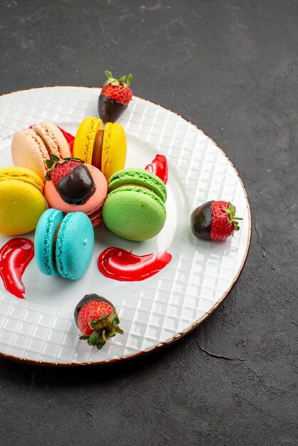 Side view from afar colorful macaroons plate of blue yellow pink and green macaroons sauce and appetizing chocolate-covered strawberries on the dark table