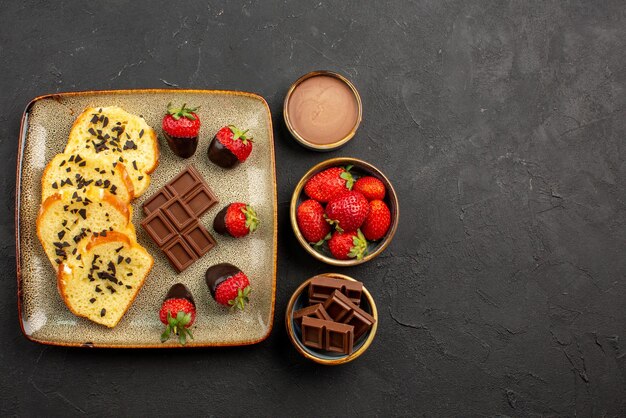 暗いテーブルのボウルにレモンとチョコレートクリームとイチゴが入ったお茶の横にあるチョコレートで覆われたイチゴとケーキの灰色のプレートと遠くのお茶の側面図