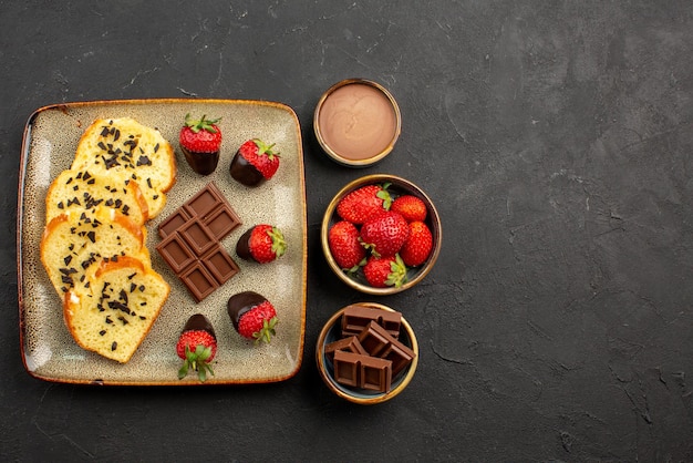 Side view from afar cup of tea with cake grey plate of cake with chocolate-covered strawberries next to the cup of tea with lemon and chocolate cream and strawberries in bowls on the dark table