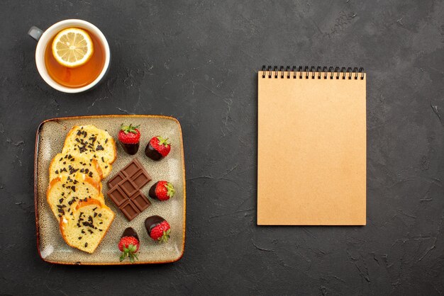 Side view from afar cup of tea with cake grey plate of cake with chocolate-covered strawberries next to the cup of tea with lemon and cream notebook on the dark table