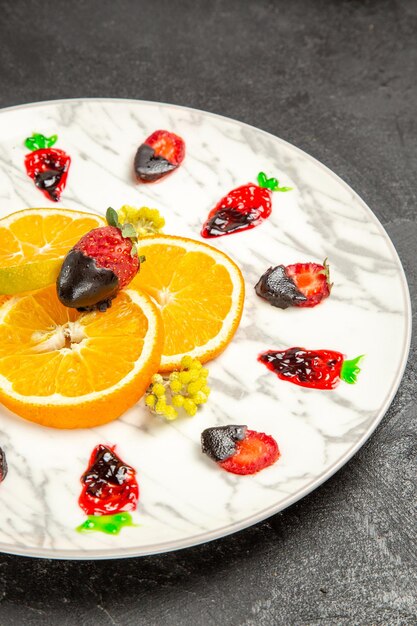 Вид сбоку издалека фрукты на тарелке дольки апельсина с клубникой в шоколаде на белой тарелке на темном столе