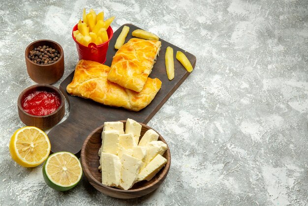 회색 테이블에 있는 치즈 케첩과 검은 후추 레몬 그릇 옆에 있는 커팅 보드에 있는 멀리 있는 파이와 케첩 식욕을 돋우는 파이와 감자튀김의 측면