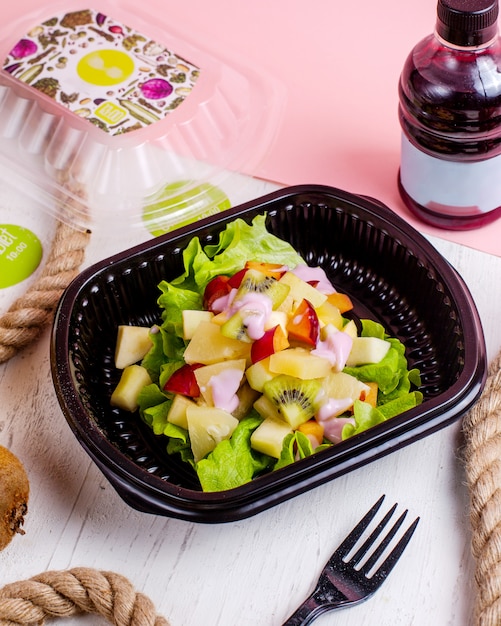 Side view of fruit salad with kivi apples pineapples and plums served with yogurt in a delivery box