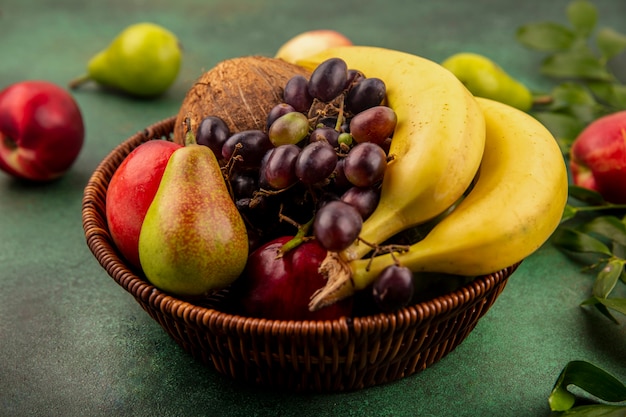 Side view of fruits as coconut banana grape pear peach in basket with leaves on green background