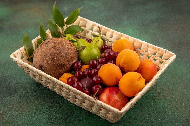 Side view of fruits as coconut peach apricot pear cherry with leaves in basket on green background