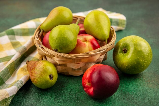 Side view of fruits as peach and pear in basket with apple and plaid cloth on green background