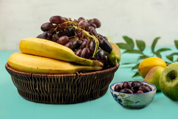 青い表面と白い背景に葉を持つブドウの果実のバスケットとリンゴレモンボウルの梨バナナブドウとしての果物の側面図