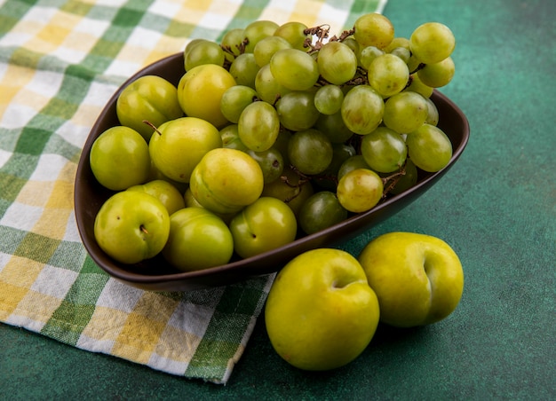 Side view of fruits as plums and grape in bowl on plaid cloth with green pluots on green background