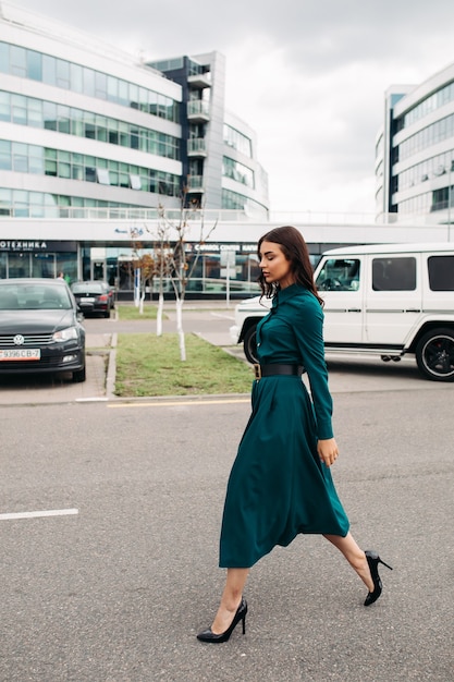現代の都市の建物に対して道路に沿って歩いている黒帯と黒のかかとと濃い緑色のドレスを着たゴージャスなブルネットのモデルの側面図のフルレングスのストックフォト。