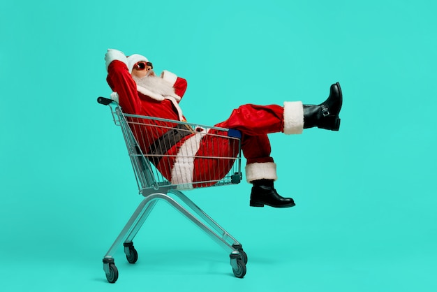 Free photo side view of funny santa claus in black sunglasses and costume making faces. old man having fun, sitting and relaxing in shopping cart