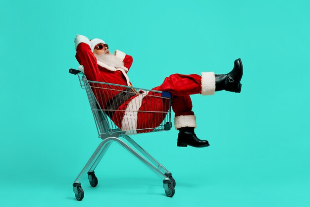 Side view of funny Santa Claus in black sunglasses and costume making faces. Old man having fun, sitting and relaxing in shopping cart 