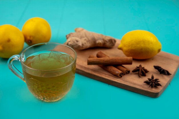 Side view of ginger juice in glass and spices as ginger and cinnamon with lemon on cutting board on blue background