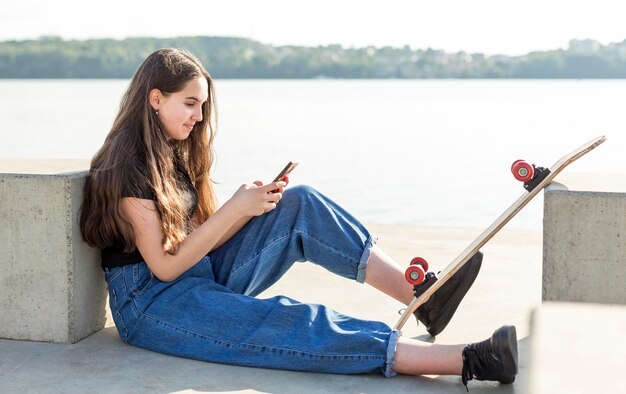 Side view girl checking her phone next to her skateboard