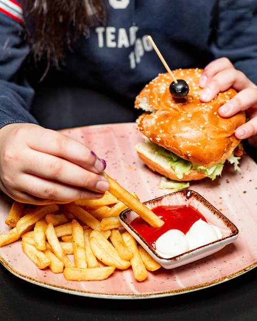 Side view of a girl eating chicken burger served with french fries and sauces at the table