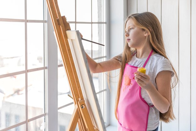 Side view of a girl holding yellow paint bottle in hand painting on the easel with paintbrush