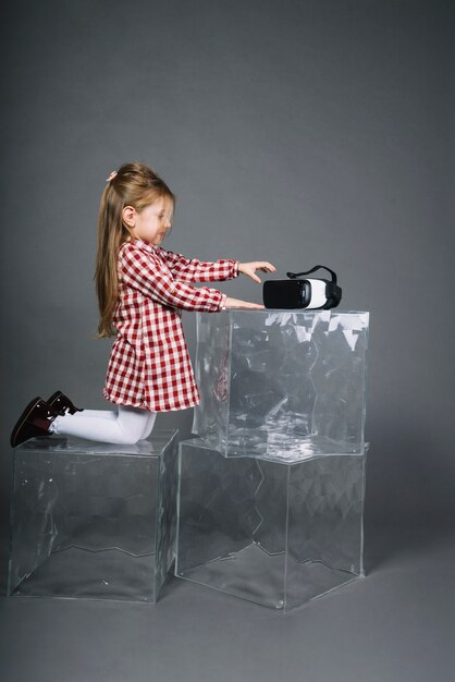 Side view of a girl kneeling on transparent cubes holding virtual reality glasses against gray background