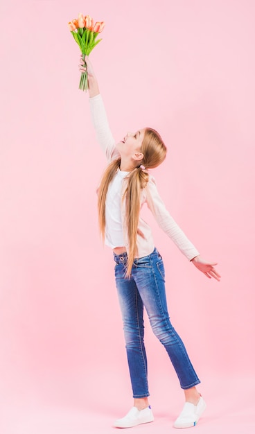 Side view of a girl raising her hand holding tulip flowers in hand standing against pink background