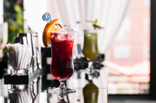 Side view of glass of cocktail with strawberry syrup rum and lemon decorated with orange slice at bar background