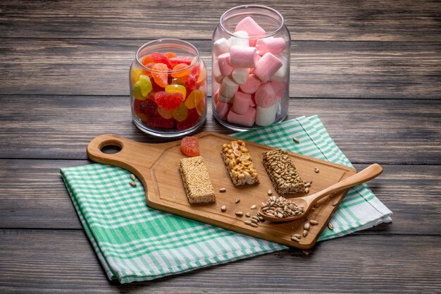 Side view of glass jars with marmalade candies and marshmallow and sweet kozinaki of sesame sunflower seeds and peanuts on a wooden board on rustic