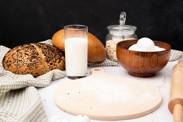 Side view of glass of milk with bowl of eggs and cutting board with cobs and oat-flakes on white surface and black background with copy space