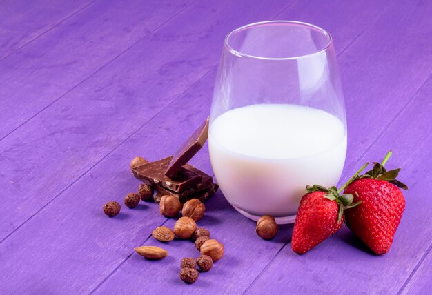 Side view of a glass of milk with fresh ripe strawberries  hazelnuts and dark chocolate on purple wooden background