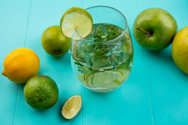 Вид сбоку на стакан воды с зеленью и лаймом с лимоном на синей поверхности