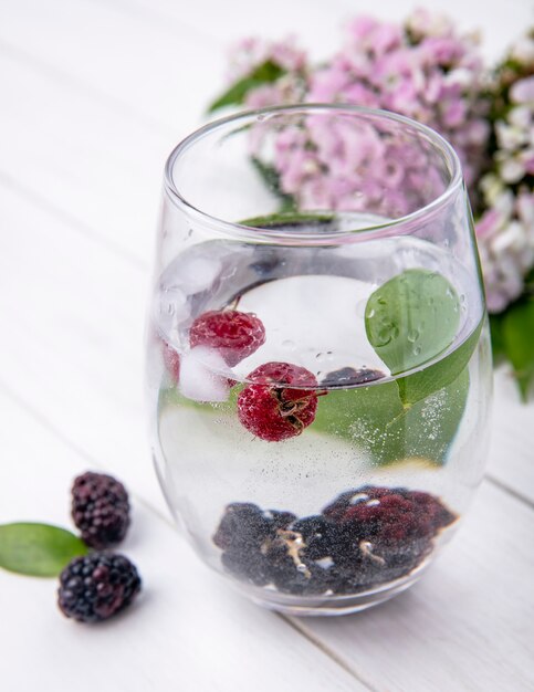 Side view of glass of water with raspberries and blackberries on a white surface