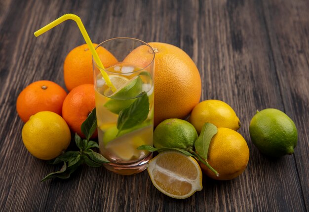 Боковой вид стакан воды с ломтиками лимона, лайма и мяты с желтой соломкой и грейпфрутом с апельсинами на деревянном фоне