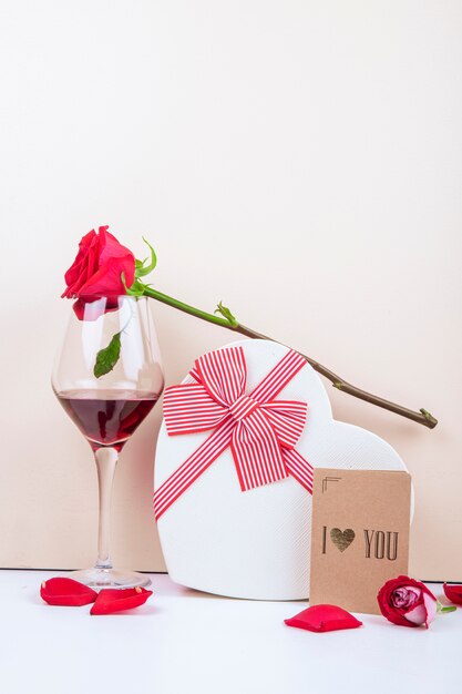 Side view of a glass of wine red color rose and a heart shaped gift box tied with bow with small postcard on white background