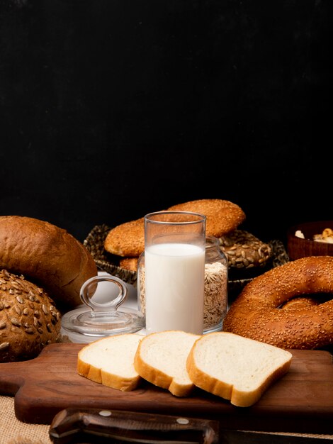 Side view of glass with milk and sliced white bread on cutting board with knife and different breads on black background with copy space