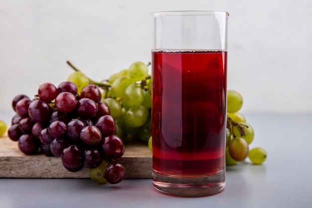Side view of grape juice in glass and grapes on cutting board on gray surface and white background