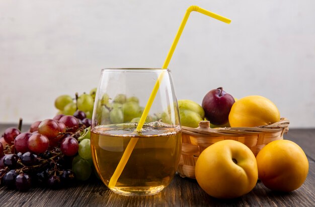 Side view of grape juice in glass and pluots nectacot in basket with grapes and nectacots on wooden surface and white background