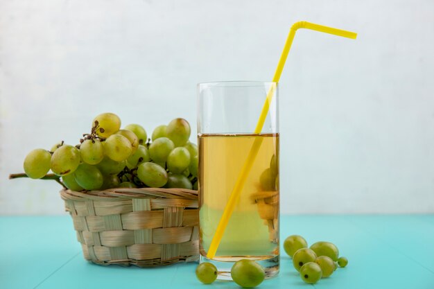 Side view of grape juice with drinking tube in glass and basket of grape with grape berries on blue surface and white background