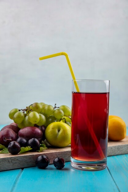 Side view of grape juice with drinking tube in glass and nectacot pluots grape on cutting board on blue surface and white background