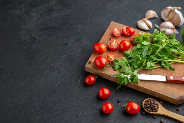 Side view of green bundle fresh whole cut tomatoes on wooden cutting board knife garlics peppers on black distressed surface