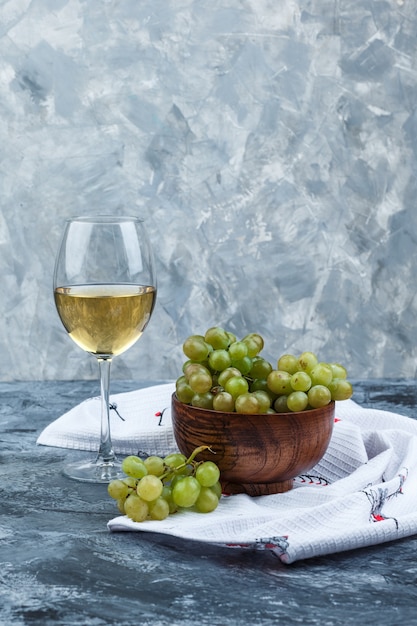 Vista laterale uva verde in una ciotola con un bicchiere di vino su intonaco grungy e sfondo di asciugatutto da cucina. verticale