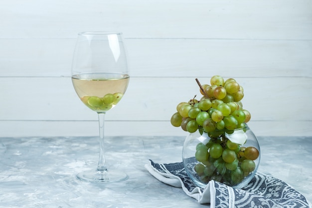 Side view green grapes in glass pot with a glass of wine, kitchen towel on wooden and grungy grey background. horizontal