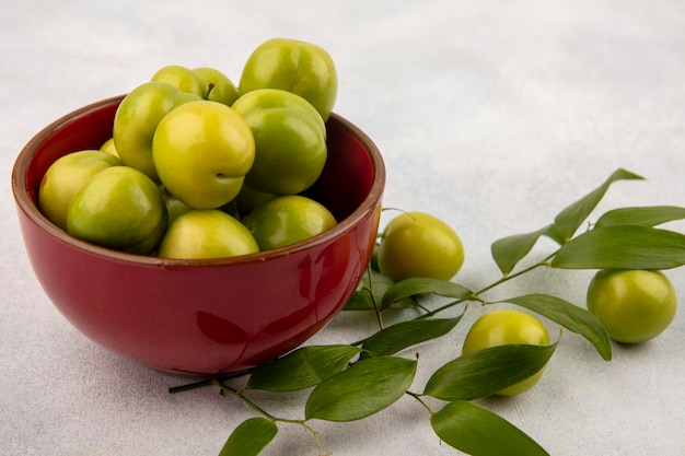 Side view of green plums in bowl with leaves on white background