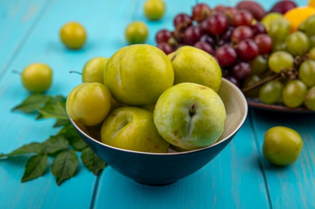 Side view of green pluots in bowl with plate of grapes plums nectacot on blue background decorated with leaves