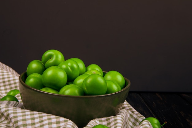 Side view of green sour plums in a bowl on plaid fabric at dark table