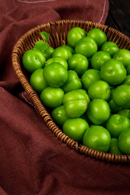 Side view of green sour plums in a wicker basket on dark red fabric table