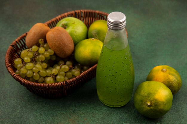 Side view green tangerines with apple kiwi and grapes in a basket with a bottle of juice on a green wall